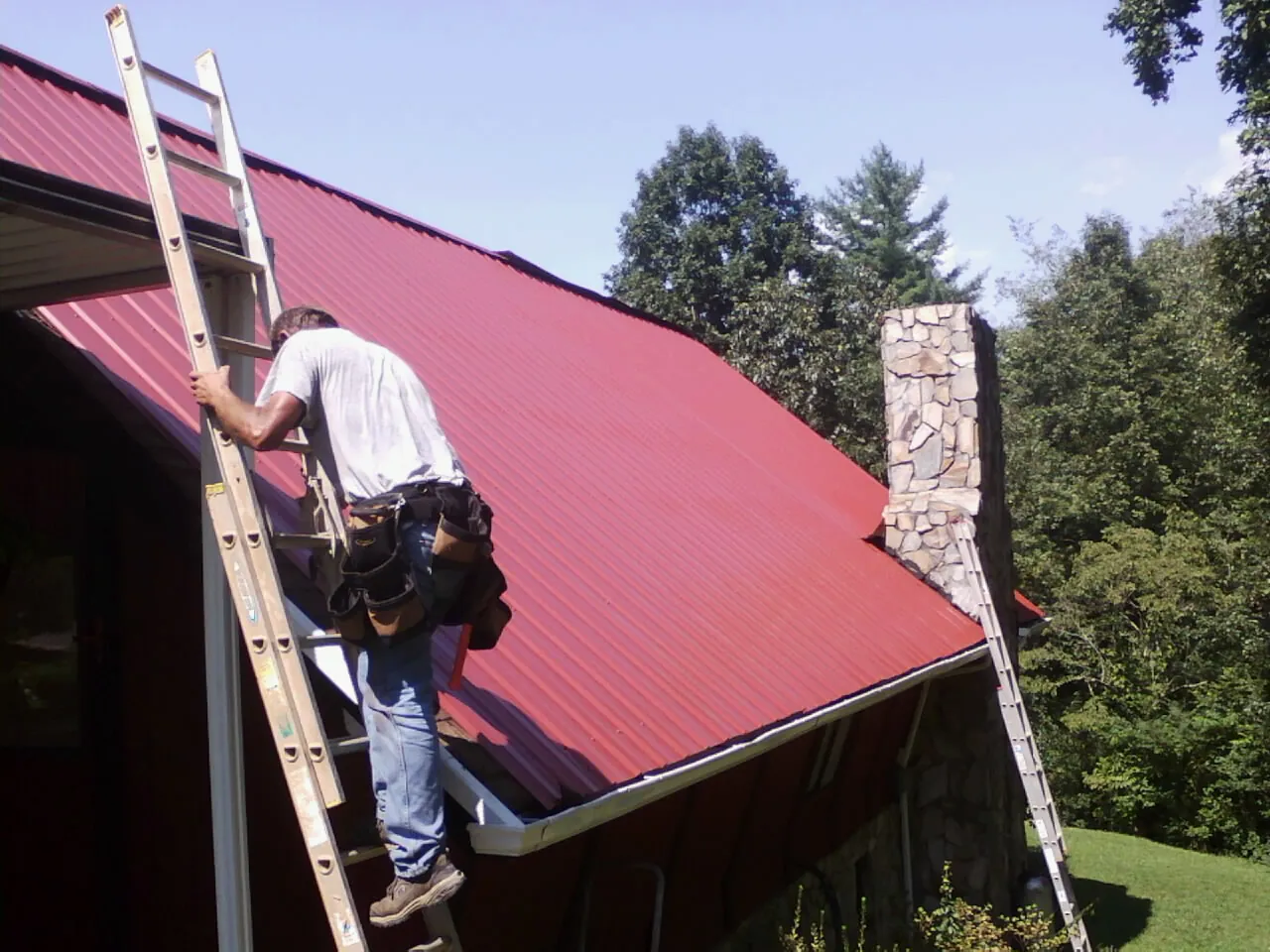 Roofer on a ladder beside a red metal roof and stone chimney — Soffit & Fascia Repair by Roofing Brothers in Callicoon